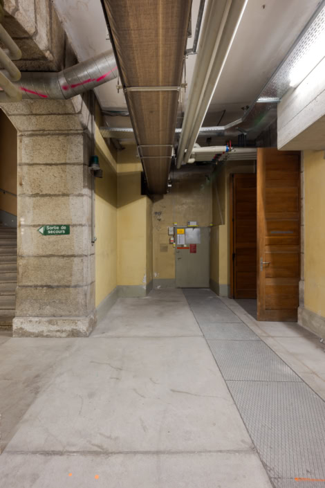 Tuyau en métal suspendu dans un sous-sol industriel, décor mural beige, porte en bois.