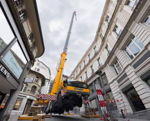Grue jaune levant une charge dans une rue étroite avec des bâtiments en arrière-plan.
