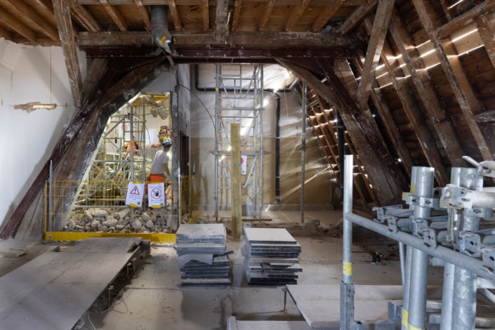 Ouvriers en rénovation dans un espace sous toiture avec charpente en bois apparente.