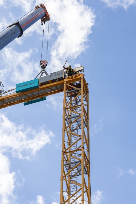 Travail en hauteur sur grue de construction avec ciel bleu et nuages blancs.