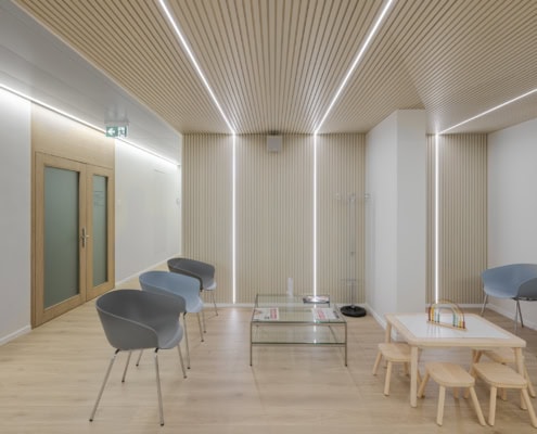 Espace d'attente avec des chaises grises, une table en verre et un coin enfant.