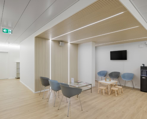 Salle d'attente avec des chaises grises, une table basse et un écran de télévision sur le mur.