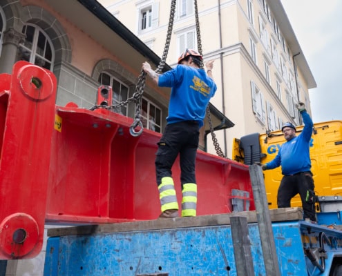 Deux ouvriers en bleu manipulant une grande pièce de construction rouge à l'aide de chaînes sur un camion.