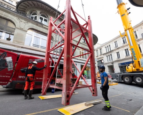 Une grue jaune soulève une structure de levage rouge sur un chantier urbain avec des travailleurs à proximité.