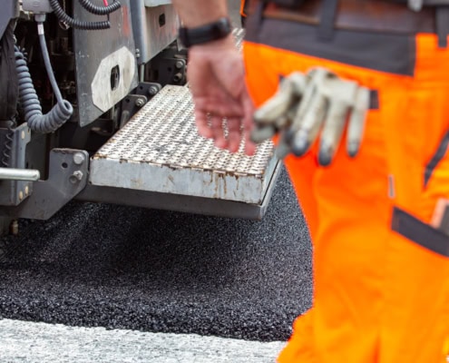 Un ouvrier en pantalon orange et gants près d'une machine de pavage sur une route nouvellement asphaltée.