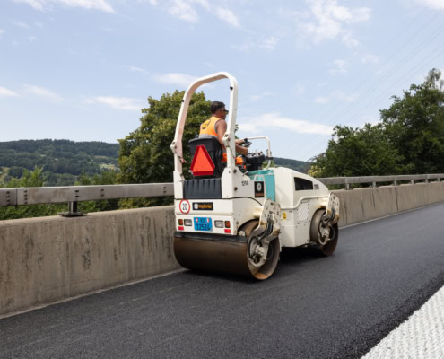 Un compacteur de route blanc et bleu en train de rouler sur une surface asphaltée fraîchement appliquée.