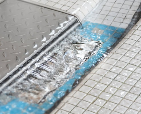 Bord d'une piscine avec un drain en métal et de l'eau qui s'écoule sur des carreaux blancs.