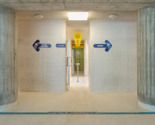 Couloir intérieur avec panneaux directionnels bleus indiquant 'Homme' et 'Femme' sur des murs carrelés.