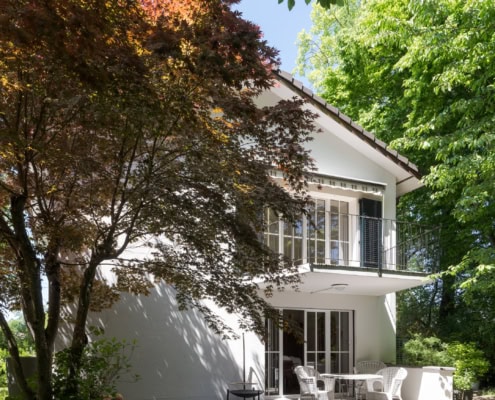 Maison blanche à deux étages avec balcon et jardin verdoyant entouré d'arbres.