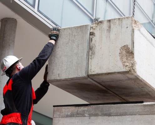 Ouvrier en gants et casque soulevant un bloc de béton à l'aide d'une grue sur un chantier.
