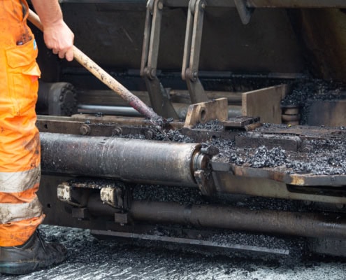 Ouvrier en combinaison orange utilisant une pelle pour appliquer de l'asphalte sur une surface routière.