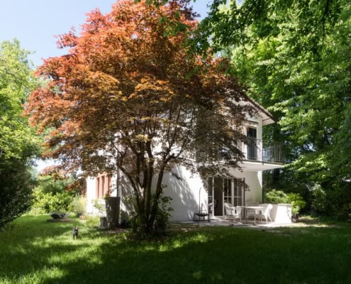 Maison blanche avec un arbre à feuilles rouges dans le jardin, entourée de verdure.