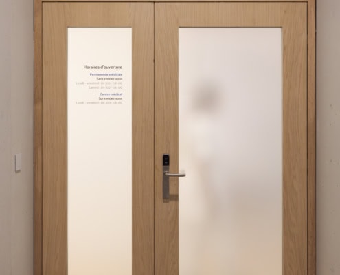 Porte en bois avec le logo Medbase et des panneaux en verre dépoli indiquant les horaires d'ouverture.