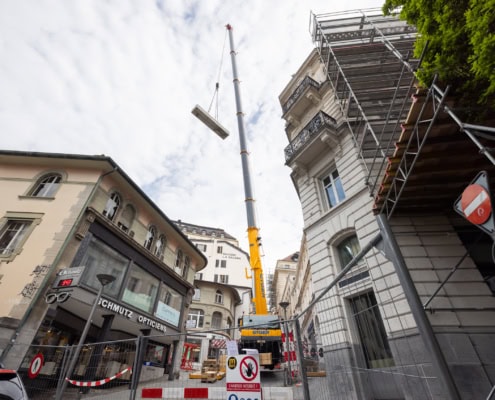 Grue jaune soulevant un panneau de construction au-dessus d'un bâtiment en rénovation dans une rue urbaine.