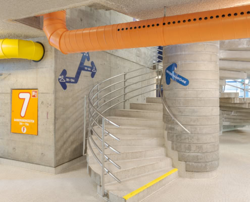 Escalier en colimaçon avec rampe en métal dans un parking souterrain, avec des tuyaux colorés au plafond.