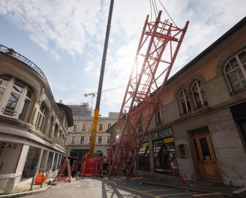 Grue rouge de construction érigée dans une rue avec des bâtiments en pierre et un ciel partiellement nuageux.