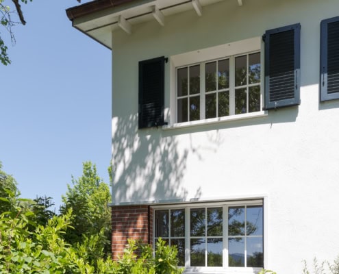 Maison à deux étages avec fenêtres à volets et végétation environnante.