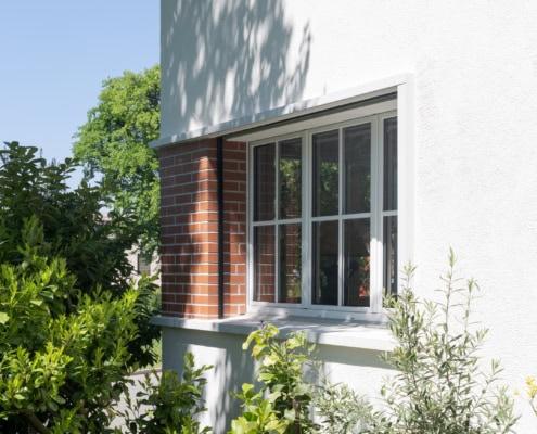 Fenêtre en bois avec des volets sur un mur blanc, entourée de verdure et d'une plante en pot.
