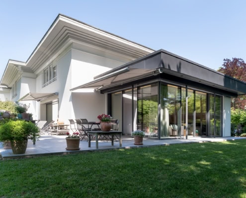 Maison moderne avec une véranda en verre et des chaises sur une terrasse extérieure.