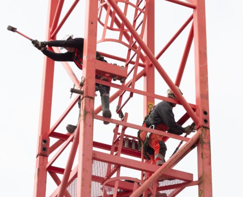 Deux ouvriers travaillant sur une grue rouge, l'un utilisant un marteau et l'autre ajustant une pièce.