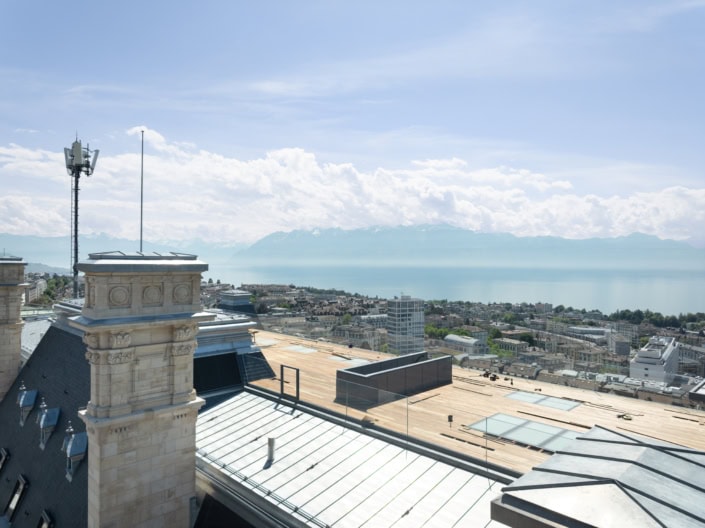Vue panoramique de Lausanne avec bâtiments et lac en arrière-plan, ciel ensoleillé.