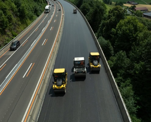 Trois rouleaux compresseurs travaillant sur une route en cours de revêtement, avec des véhicules en circulation.