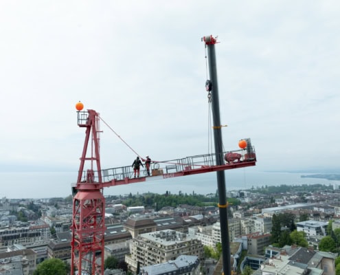 Travailleurs sur une grue rouge surplombant une ville avec un lac en arrière-plan.