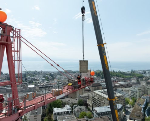 Grue rouge en train de soulever un bloc de béton au-dessus de la ville de Lausanne avec vue sur le lac.