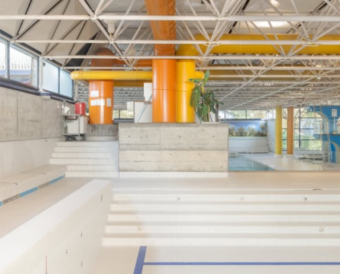 Intérieur d'une piscine montrant des escaliers en béton et des structures métalliques colorées au plafond.