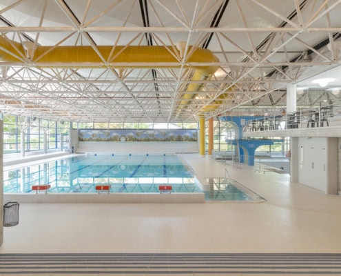Intérieur d'une piscine avec gradins, horloge et accès aux vestiaires visibles en arrière-plan.