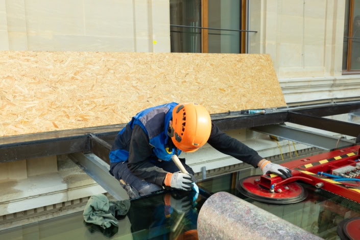 Travailleur en construction avec casque orange, pose de vitrage sur un bâtiment ancien.